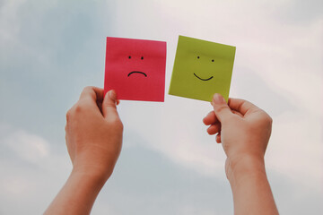 Woman hand holding paper note smile and sad face emotion on sky background.