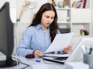 Young female secretary working in the office of a large company is studying the documentation on paper, sitting at ..desktop