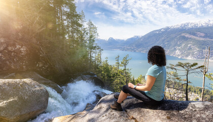 Adventurous Adult Woman is sitting on top of a Beautiful Waterfall, Shannon Falls, and watching the view. Taken near Squamish, North of Vancouver, British Columbia, Canada.