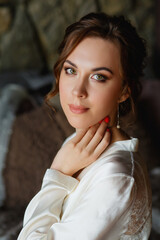 Fototapeta premium Morning of the bride. A gorgeous bride, a beautiful woman in a white coat at the window. Morning of the bride before the wedding. Natural beauty and professional makeup.