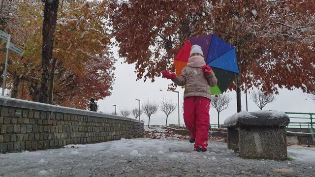 Little kid girl with colorful umbrella and scarf plays kicking snow. Slow-motion low angle