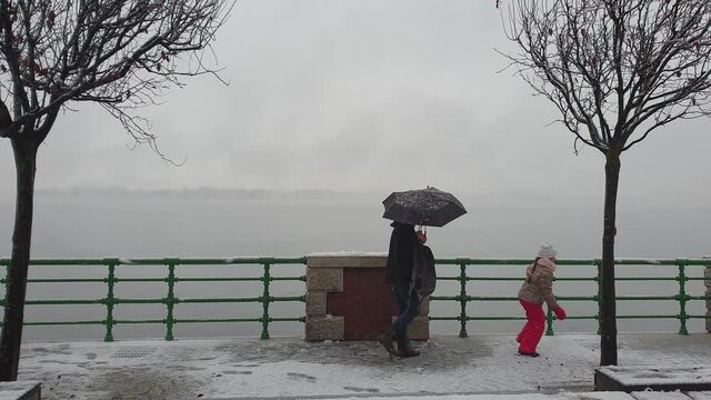 Mother with umbrellas and child with red trousers walk outdoors on snowy seafront. Slow-motion