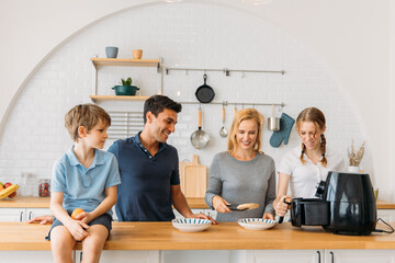 Cheerful caucasian handsome father and beautiful mother with son and daughter preparing healthy snacks in kitchen using air fryer at home - 430513653