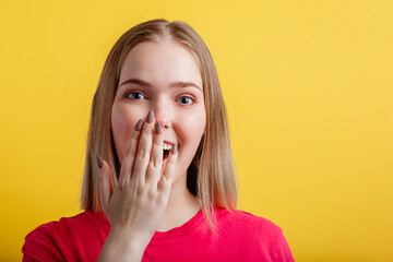 Young woman is pleasantly surprised. Teenage blonde girl covered her open mouth with hand in surprise. Positive emotional portrait of young woman on color yellow background with copy space.