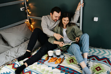 Immersed middle aged couple having a breakfast in a bed and reading a book