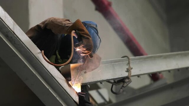 A welder wearing a safety mask, safety helmet, welding goggles. Professional welder welding steel structure with spark in factory.