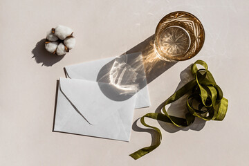 Still life scene with cotton flower on beige background in sunlight and blank envelope with ribbon, invitation mockup.