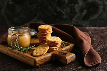 Board with tasty crackers and jar with peanut butter on dark background