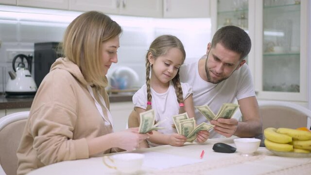 Joyful Caucasian young family scattering money in slow motion resting in kitchen at home. Portrait of positive father mother and daughter enjoying wealth indoors. Richness and lifestyle