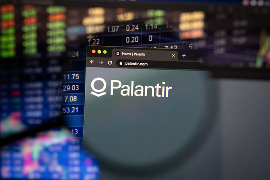 Palantir company logo on a website with blurry stock market developments in the background, seen on a computer screen through a magnifying glass