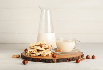 Cup with hot white chocolate and jug of milk on light wooden background