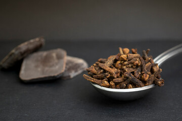tea spoon full of cloves, with dark chocolate tablets blurred in the background, isolated on black