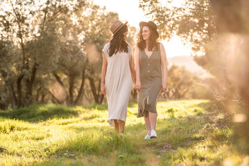 Fototapeta premium two friends, a lesbian couple with hats on their heads walking hand in hand in the field on a sunny day. lesbian concept