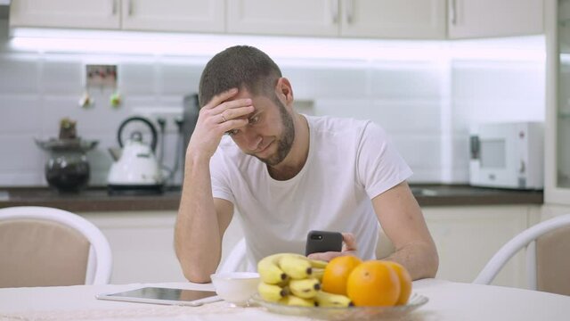 Stressed handsome Caucasian man thinking and holding head in hands sitting in kitchen at home. Portrait of worried anxious guy having problems indoors. Anxiety and stress concept