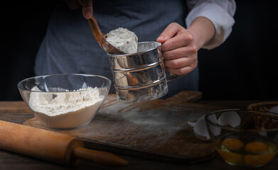 Women's hands, flour and dough. A woman in an apron prepares dough for homemade baking, a rustic home cozy atmosphere, a dark background with unusual lighting.
