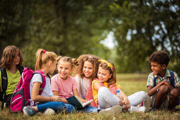 Fototapeta premium Large group of school kids having fun in nature. Little girl reading book to her friends.