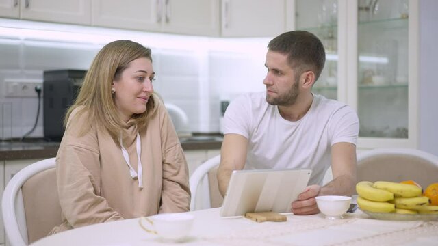Relaxed young Caucasian couple surfing Internet on tablet and talking at home. Portrait of man and woman resting on weekend in kitchen indoors. Lifestyle and family concept. Slow motion