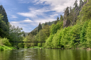 View of the Svatosh Rocks (Czech Republic, Karlovy Vary) on a sunny spring day. High natural rocks surrounded by bright green young greenery and a suspension bridge over the river. Blue-white sky 