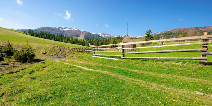mountainous rural landscape in spring. haystack on a grassy field behind the wooden fence on rolling hills. snow capped ridge in the distance. beautiful countryside scenery on a bright sunny day