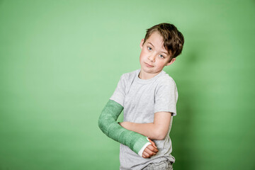 cool young schoolboy with broken arm and green plaster posing in front of green background in the studio