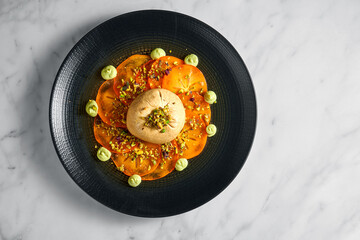 Buratta cheese with persimmon and pistachio in a black plate on a light marble background