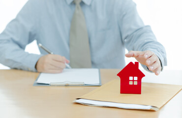 A House on document envelope with man signing purchase documents in background. while hand complete the insurance policy, rental documents and loan document. concept guy buying new house