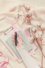 Women hand holding cosmetic on beige draped fabric with cotton branches and notebooks.  Top view. Modern aesthetic flat lay .