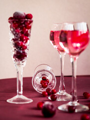 Fototapeta na wymiar red berries scattering from a crystal glass lying on the table among glasses with pink alcoholic drink, many cranberries and cherries on crystal wine glass