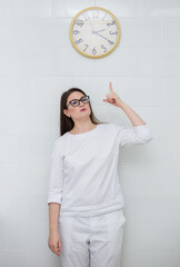 a female doctor wearing glasses and a white medical uniform stands and points at her watch
