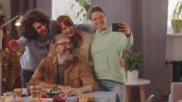 Cheerful mother, father and teenage children smiling and posing together for smartphone camera while taking selfie at home dinner