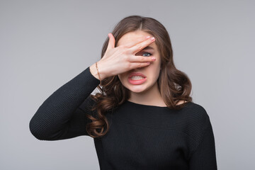 Worried, scared young woman looking between fingers, isolated over gray background. Half length...