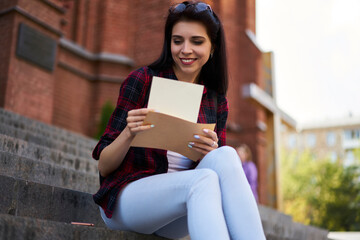 Fototapeta premium Happy millennial student with yellow envelope reading received mail notification during daytime in city, Caucasian hipster girl checking message and correspondence holding postal paper and postcard