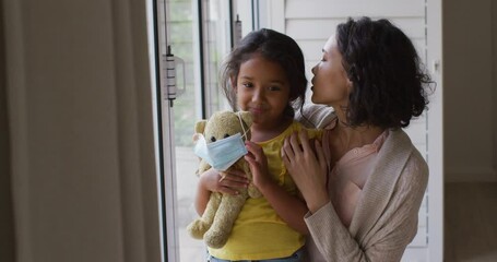 Hispanic mother and daughter embracing in window holding teddy bear with face mask - Powered by Adobe
