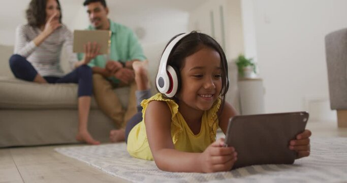 Happy hispanic girl watching video on tablet with parents sitting on sofa in background