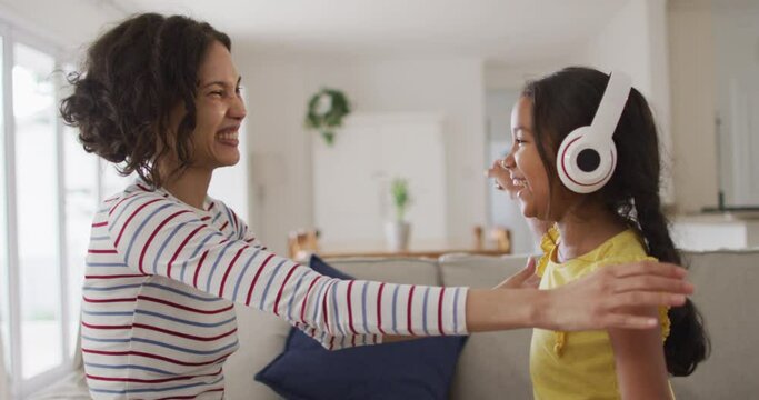 Happy hispanic mother and daughter wearing headphones listening to music sitting on sofa