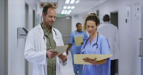 Caucasian male and female doctor talking in hospital corridor looking at tablet and patient files