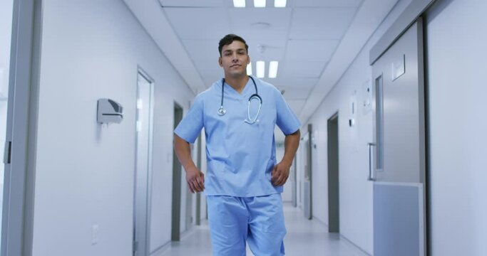 Portrait of smiling mixed race male doctor wearing scrubs walking in hospital corridor
