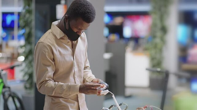 young black guy is shopping in electronics store during pandemic, wearing mask on face, choosing smartwatch