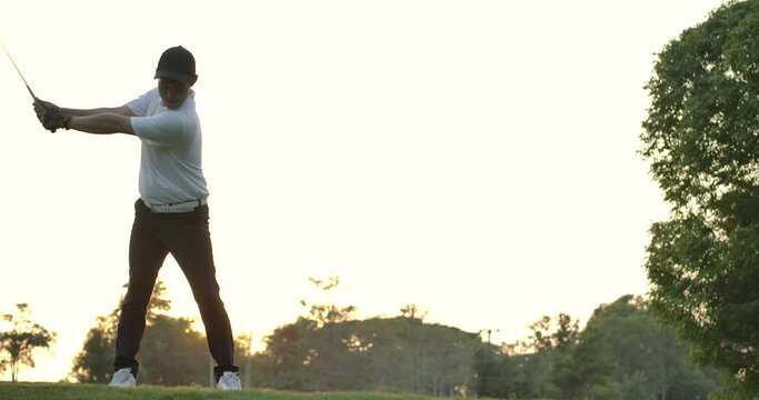 Asian male golfer swinging golf club and hitting golf ball with sunset background in slow motion.