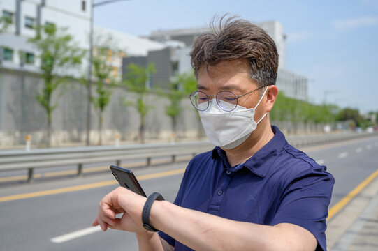Middle-aged Asian man wearing a face mask and looking at a smartphone in the driveway.