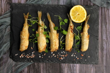 fried breaded fish served on a black stone, decorated with fresh lemon, dark wooden background. horizontal view from above
