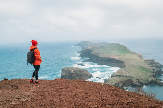  Female dressed orange sporty hoodie and red cap with backpack enjoying Atlantic ocean view on Ponta de Sao Lourenço peninsula - the easternmost point of Madeira island, Portugal