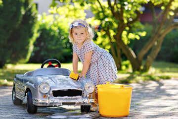 Cute gorgeous toddler girl washing big old toy car in summer garden, outdoors. Happy healthy little child cleaning car with soap and water, having fun with splashing and playing with sponge.