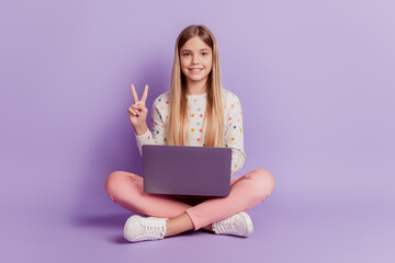 Positive kid sit legs crossed use laptop make v-sign isolated over violet background