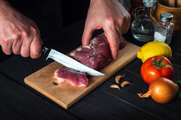 Chef cutting beef meat with knife on kitchen, cooking food. Vegetables and spices on kitchen table...