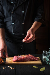 Chef sprinkles the raw beef meat with salt. Preparing meat before baking. Working environment in kitchen of restaurant or cafe