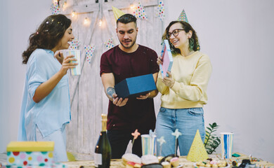 Young hipsters greeting girl on her birthday party opening decorative box with surprise during celebration, happy young woman amazed with gift from best friends satisfied with holiday and festive mood