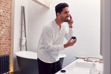 Man Wearing Pyjamas At Home In Modern Bathroom Putting On Moisturizer Looking In Mirror