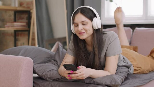 PAN shot of young woman in headphones relaxing on couch and listening to music while browsing social media on mobile phone