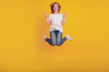 Fototapeta na wymiar Portrait of lovely friendly girl jumping show two v-signs on yellow background